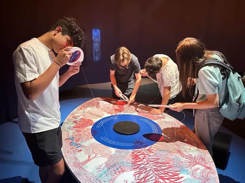 Expanish summer camp students in Barcelona exploring an interactive museum exhibit, closely examining details with magnifying tools around a colorful display table.